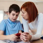 Smiling boy using smartphone with his mother at home